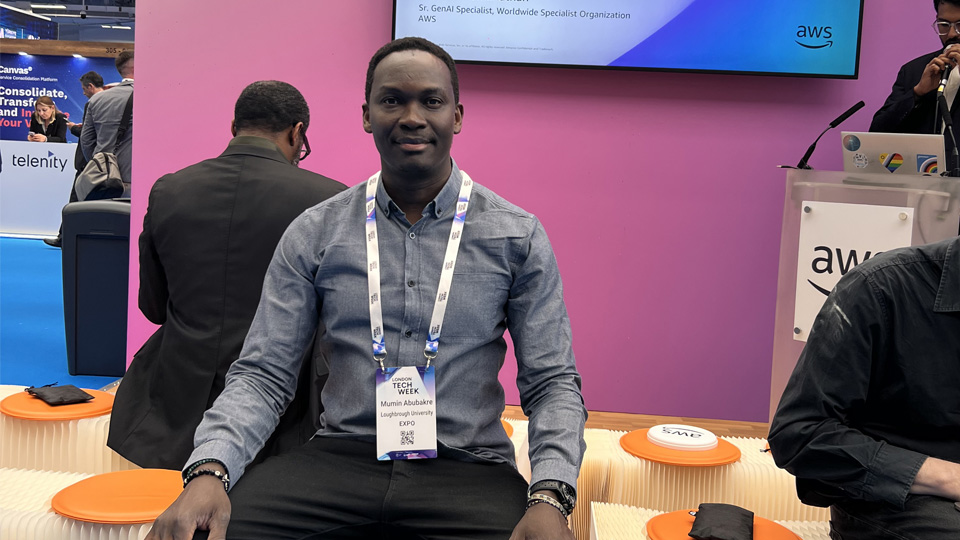 A man sits on an orange stool in front of a pink wall with a screen.